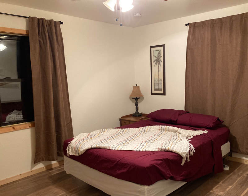 A bedroom at night with the ceiling light on: brown curtains on two windows. A large bed with dark red sheets, and a light throw blanket over the bed. In the far corner is a picture of a palm tree, a lamp, and a night stand. 