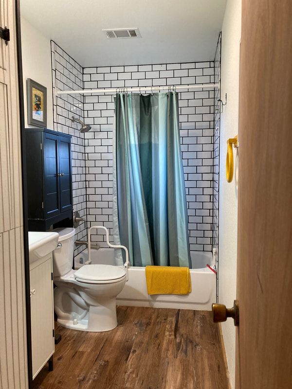 Looking through the doorway into a bathroom. The far wall is tiled with white tile. A white bathtub with a yellow bath rug and green shower curtain is against the far wall. A toilet, small black cabinet, and sink are visible. There is a lot of light. 